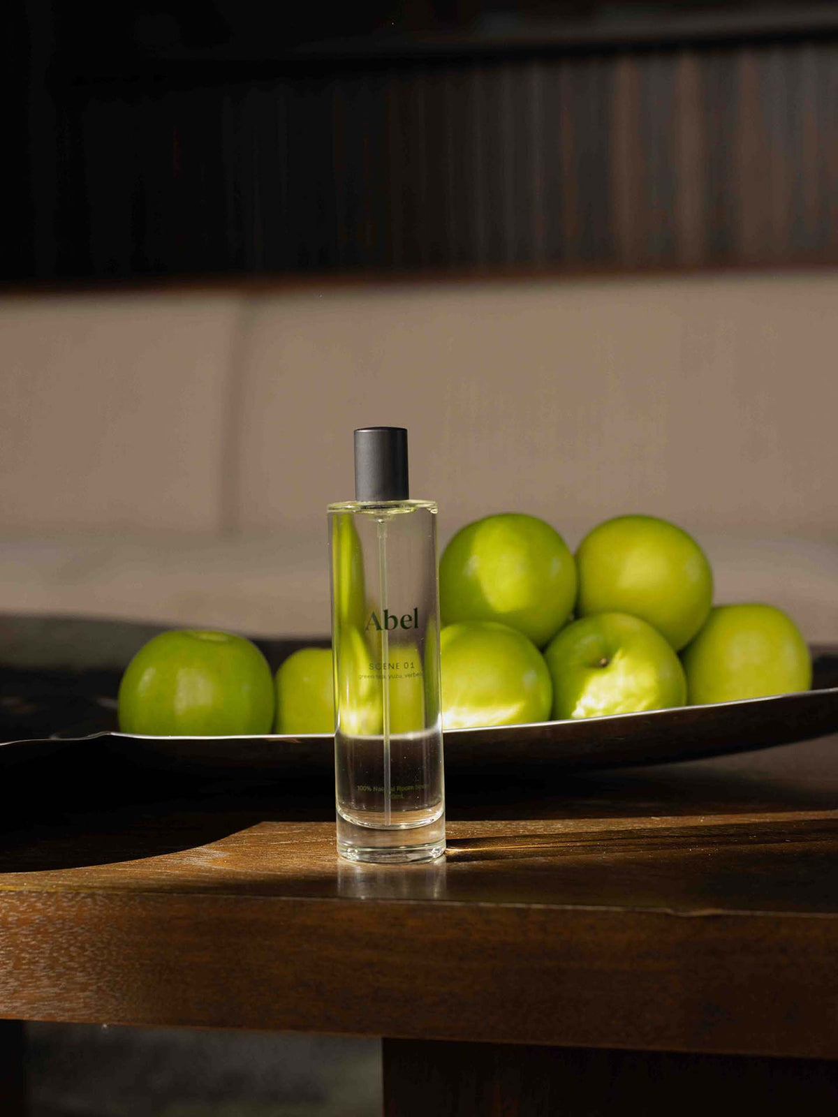 A bottle of Abel&#39;s Room Spray – Scene 01, featuring the refreshing aromas of green tea, yuzu, and verbena, stands on a wooden table beside a bowl filled with green apples.