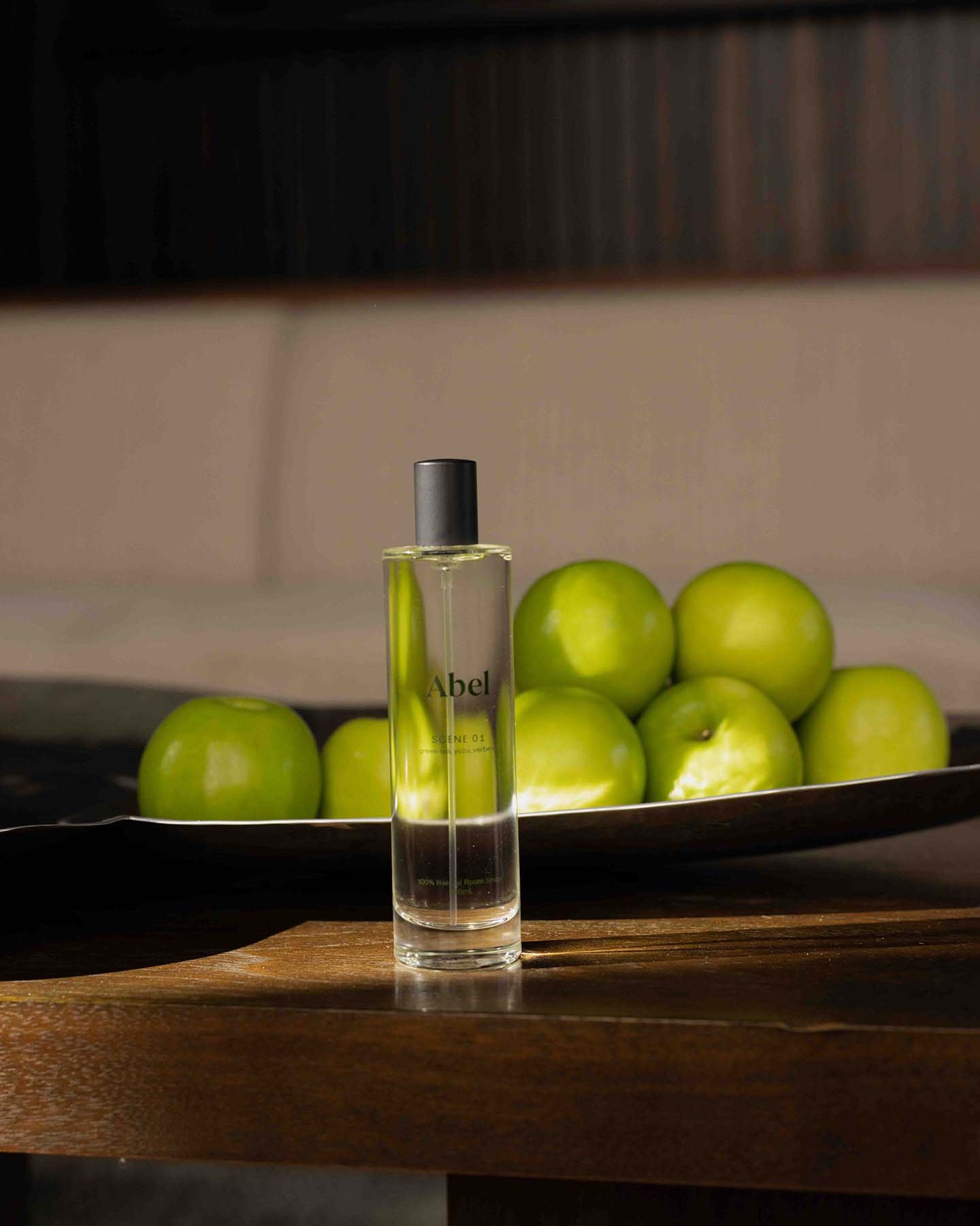 A bottle of Abel's Room Spray – Scene 01, featuring the refreshing aromas of green tea, yuzu, and verbena, stands on a wooden table beside a bowl filled with green apples.