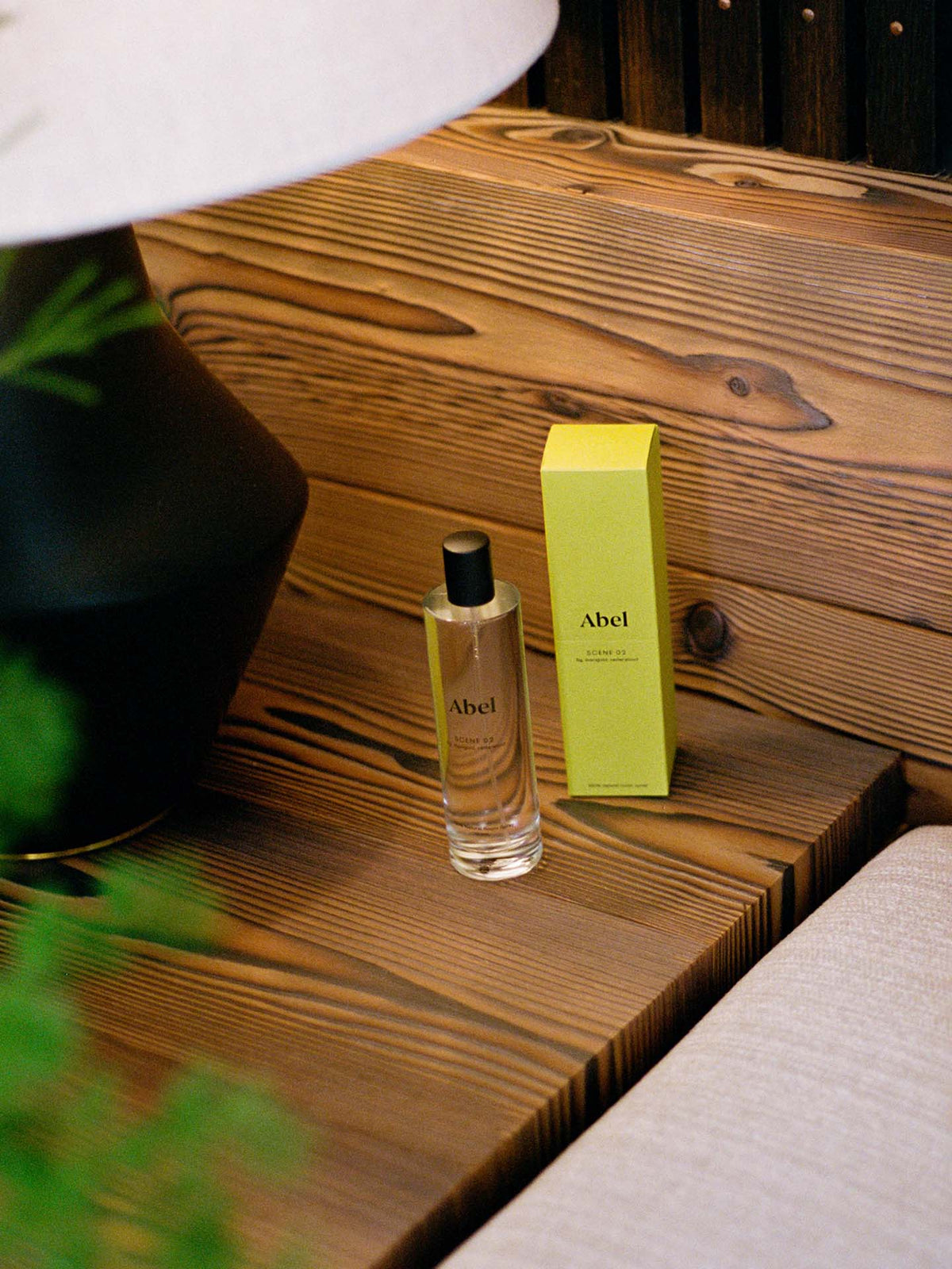 A bottle of Abel&#39;s &quot;Room Spray – Scene 02 ⋅ fig, marigold, cedarwood&quot; and its green box sit elegantly on a wooden surface beside a lamp, releasing scents of fig, marigold, and cedarwood.