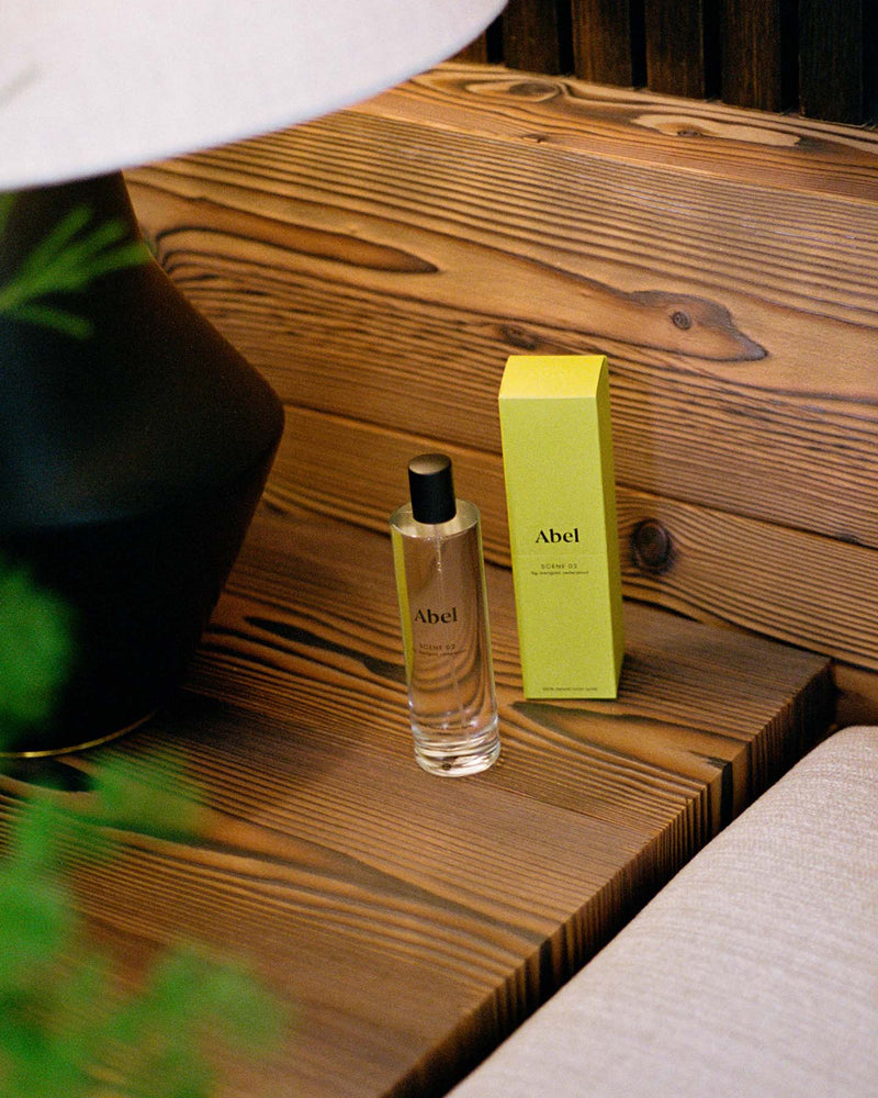 A bottle of Abel's "Room Spray – Scene 02 ⋅ fig, marigold, cedarwood" and its green box sit elegantly on a wooden surface beside a lamp, releasing scents of fig, marigold, and cedarwood.