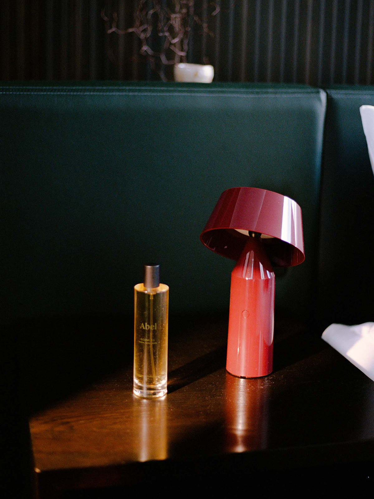 A bottle of &quot;Room Spray – Scene 03&quot; by Abel sits beside a red modern lamp on a dark wooden table, creating an atmosphere of quiet luxury against the backdrop of green cushions, embodying an elevated scent ritual with its leather, tonka, and vanilla notes.