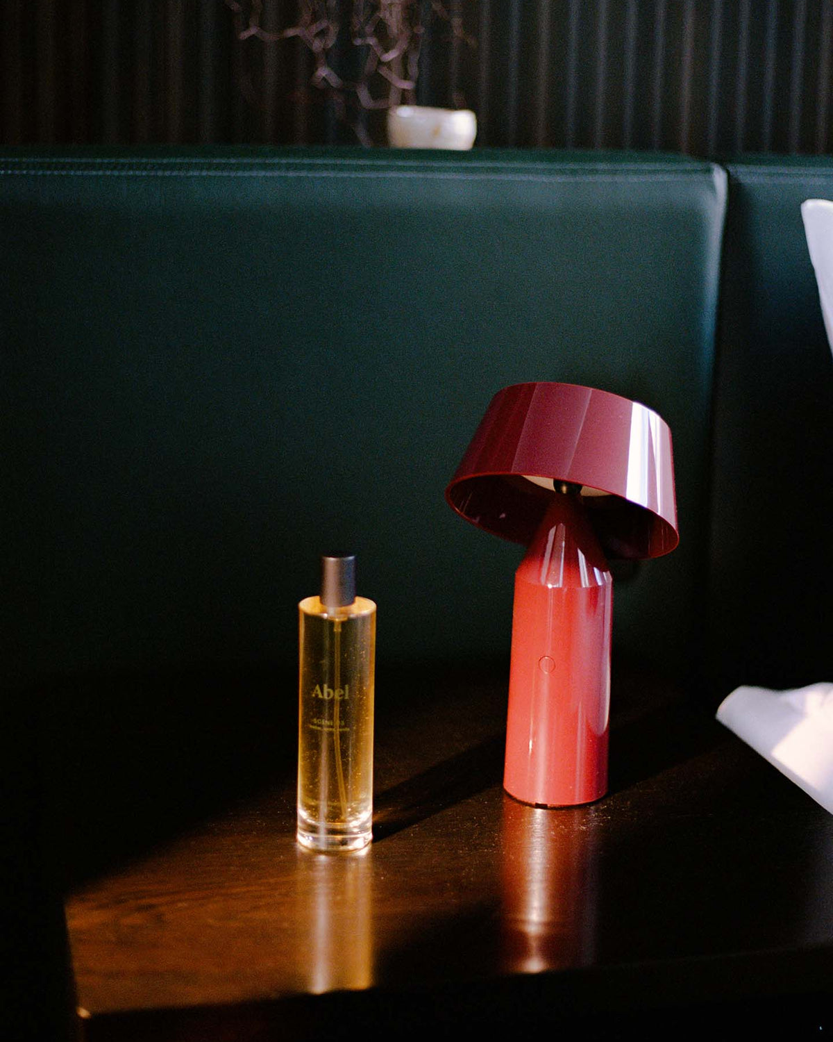 A bottle of "Room Spray – Scene 03" by Abel sits beside a red modern lamp on a dark wooden table, creating an atmosphere of quiet luxury against the backdrop of green cushions, embodying an elevated scent ritual with its leather, tonka, and vanilla notes.