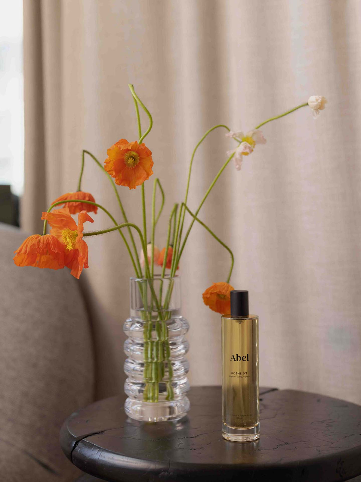 A glass vase with orange flowers elegantly adorns the round table, exuding a sense of refined luxury. Accompanying it is a bottle of Abel&#39;s Room Spray – Scene 03, which infuses the air with notes of leather, tonka, and vanilla to enhance an elevated scent ritual. The tranquil background features a curtain and a glimpse of the couch.