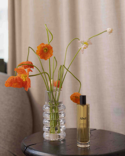 A glass vase with orange flowers elegantly adorns the round table, exuding a sense of refined luxury. Accompanying it is a bottle of Abel's Room Spray – Scene 03, which infuses the air with notes of leather, tonka, and vanilla to enhance an elevated scent ritual. The tranquil background features a curtain and a glimpse of the couch.