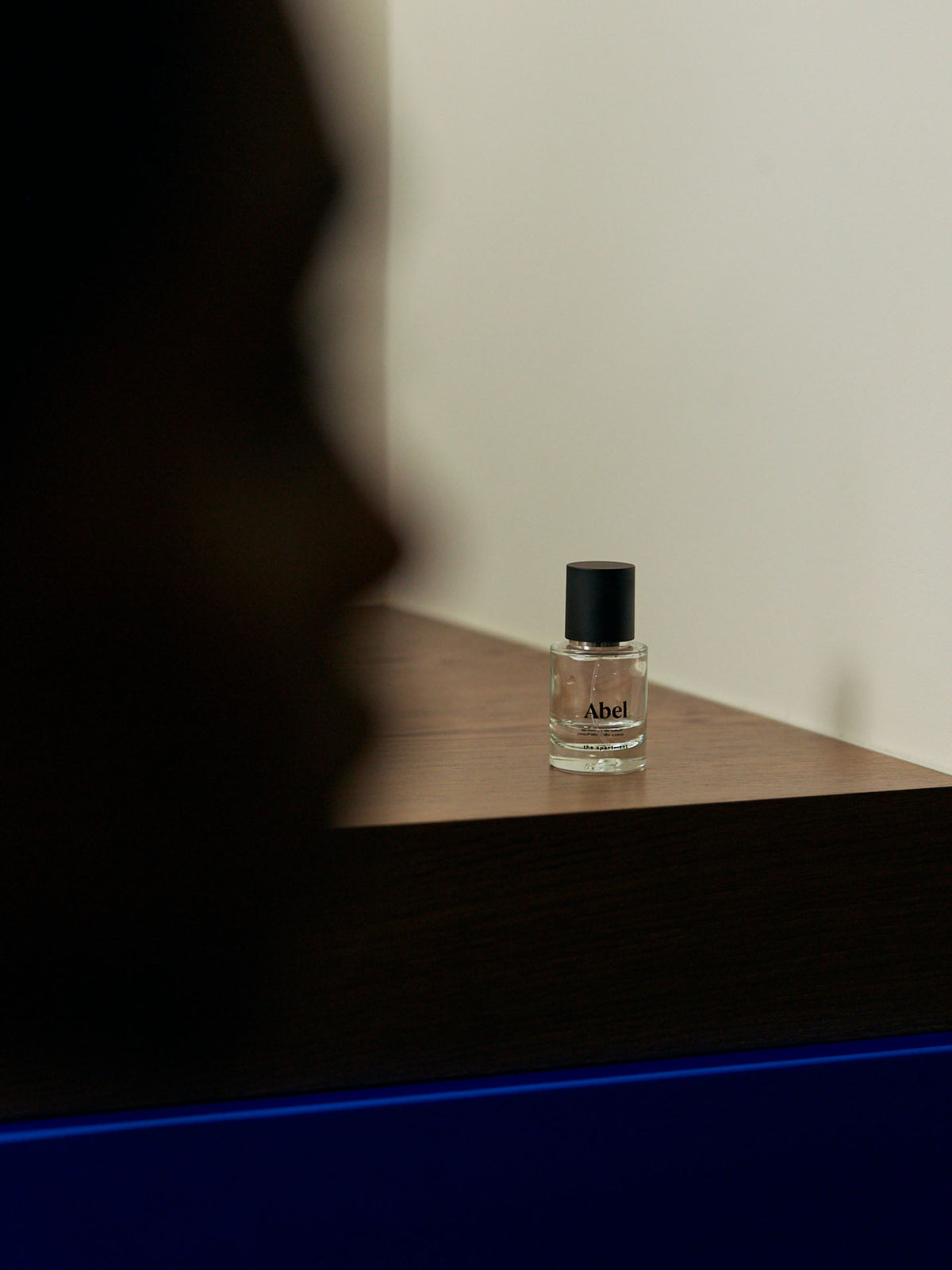 A small bottle of Abel&#39;s &quot;The Apartment&quot; Eau de Parfum rests on a wooden shelf, with a silhouetted person in the foreground, exuding an aura of dark and sophisticated gourmand elegance.