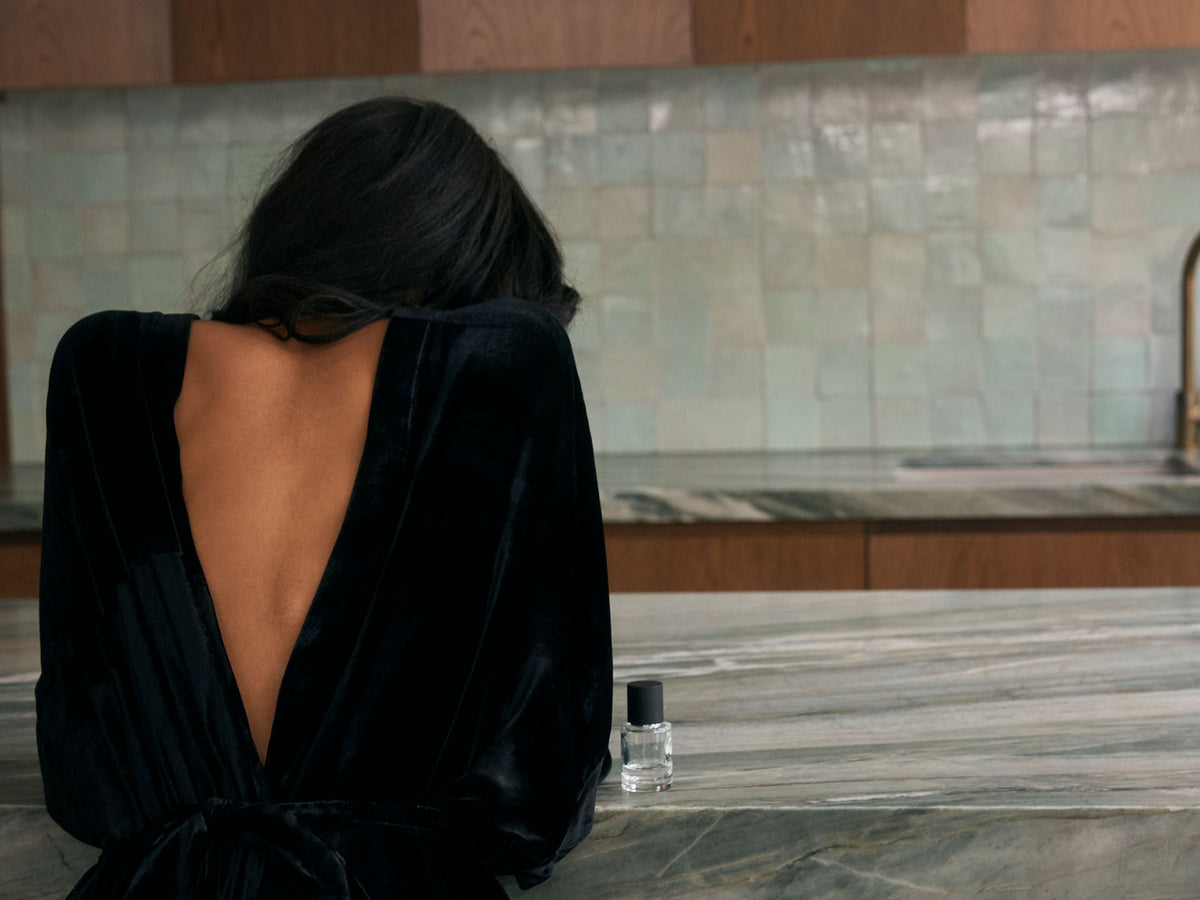 A person with long hair and a backless black dress, exuding Parisian chic, stands at a marble countertop with a small bottle of &quot;The Apartment&quot; by Abel nearby. The background features tiled walls and elegant wooden cabinets.