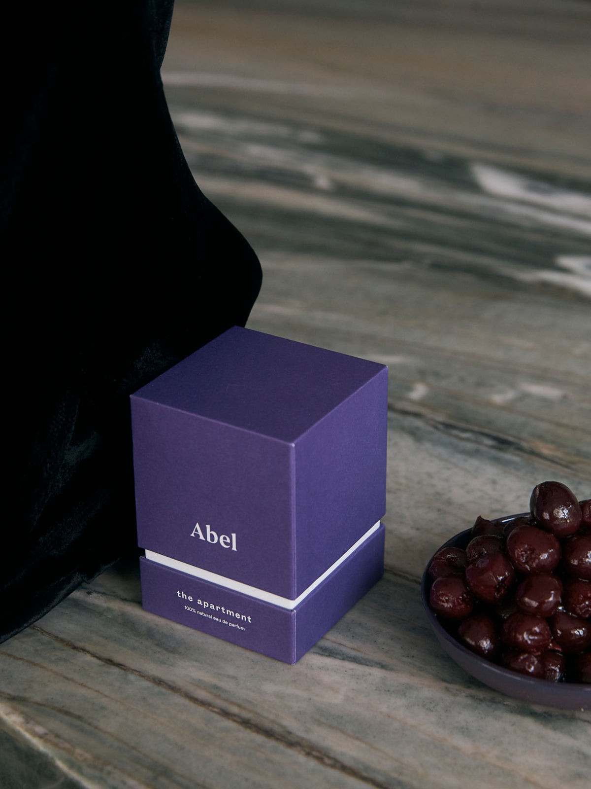 A dark and sophisticated gourmand perfume box, labeled &quot;Abel&quot; and named &quot;The Apartment,&quot; exudes Parisian chic as it sits elegantly beside a bowl of dark cherries on a marble surface, hinting at its floral gourmand essence.