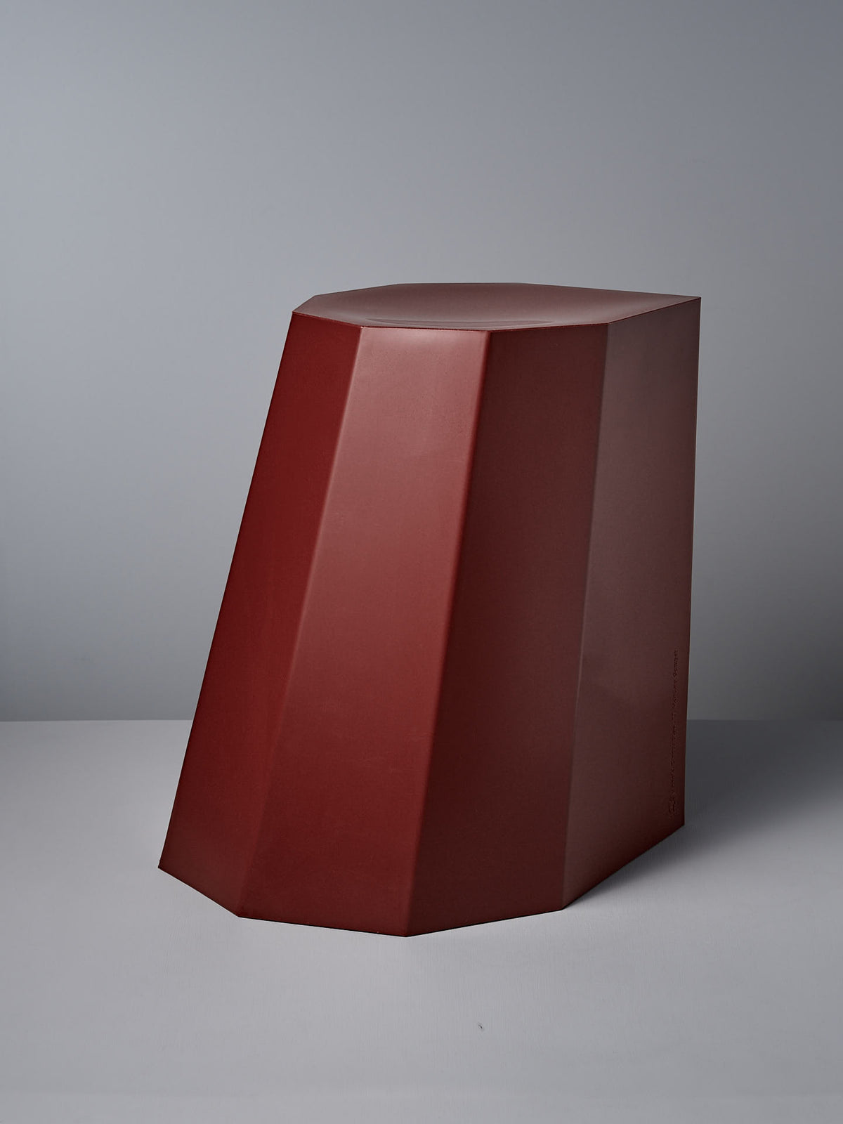 The Arnold Circus Stool by Martino Gamper, in heritage red, features a geometric design with faceted sides and is made from recyclable polyethylene, blending sustainability with modern style against a gray backdrop.