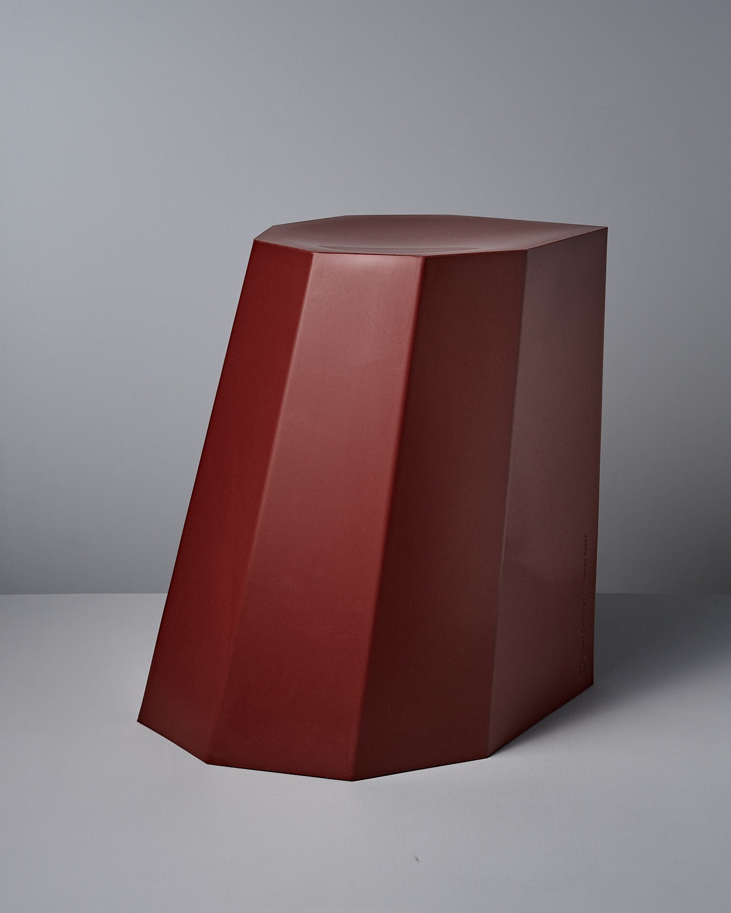 The Arnold Circus Stool by Martino Gamper, in heritage red, features a geometric design with faceted sides and is made from recyclable polyethylene, blending sustainability with modern style against a gray backdrop.