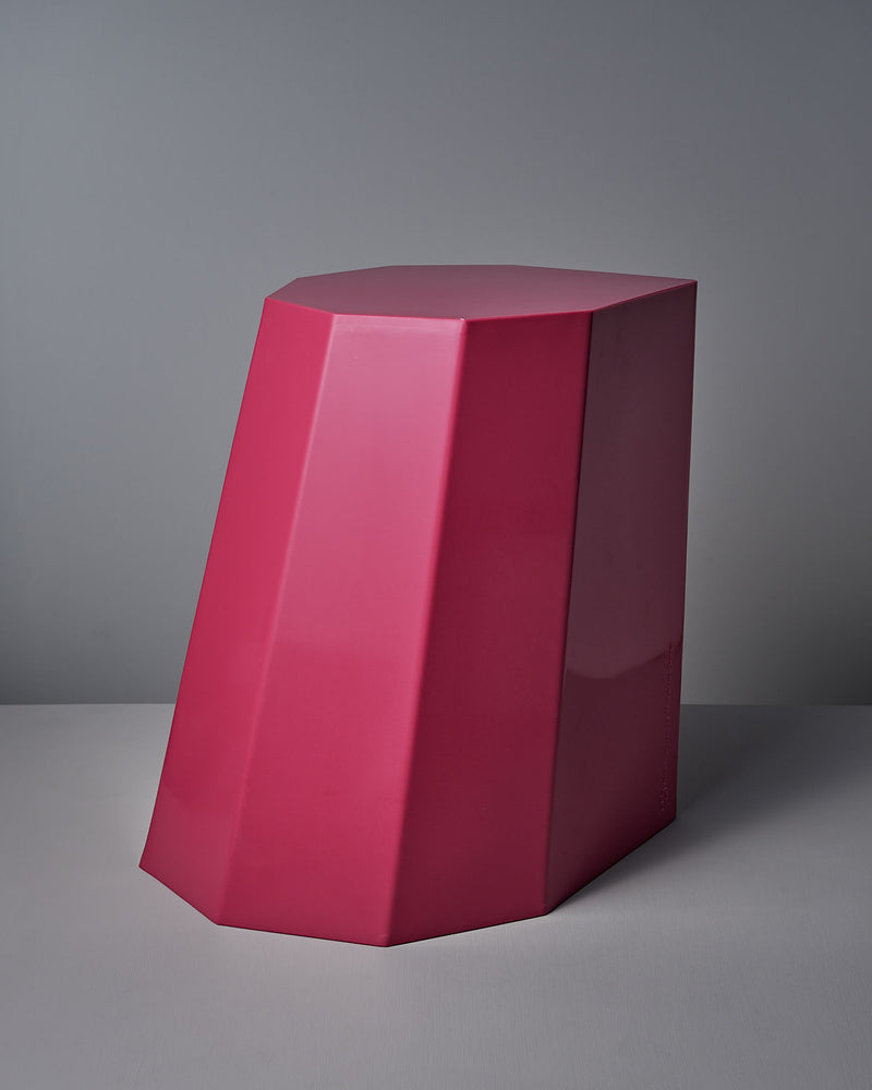 The Arnold Circus Stool in Magenta by Martino Gamper is a large, geometric sculpture crafted from recyclable polyethylene. Its angular, faceted design features smooth surfaces and is displayed against a gray backdrop.