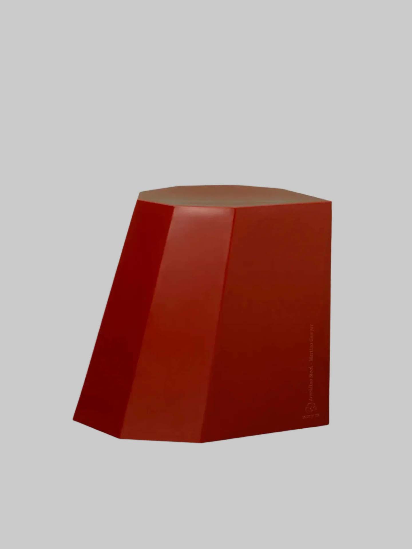 A red, angular, geometric object with a flat top and uneven sides sits on a gray background, reminiscent of the Arnoldino Stool – Heritage Red by Martino Gamper.