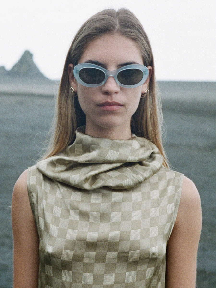 Woman with long hair wearing checkered sleeveless top and sunglasses, standing outdoors with rocks and sky in the background. The perfect eyewear sizing of the Paloma Sunglasses – Sky from auór complements her style, offering optimal lens size for both fashion and function.