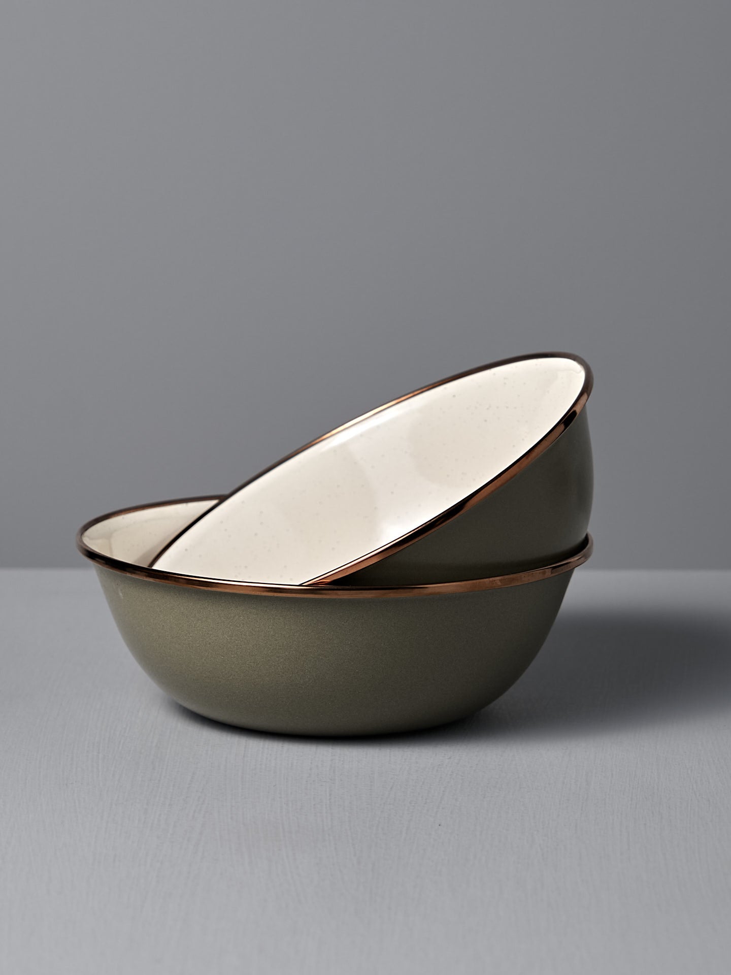 The Barebones Enamel Two-Tone Bowl – Olive Drab (Set of 2) features olive green exteriors and white interiors, shown stacked on a gray surface with a matching background.