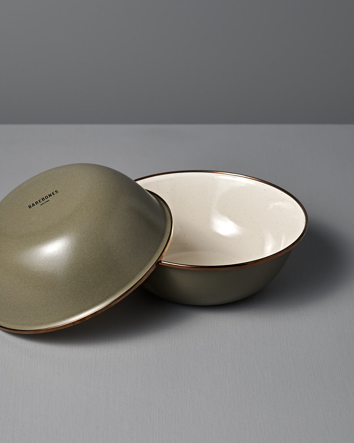 The Barebones Enamel Two-Tone Bowl – Olive Drab (Set of 2) features matte green bowls with brown rims and a vintage-inspired design, displayed on a grey surface against a grey background.