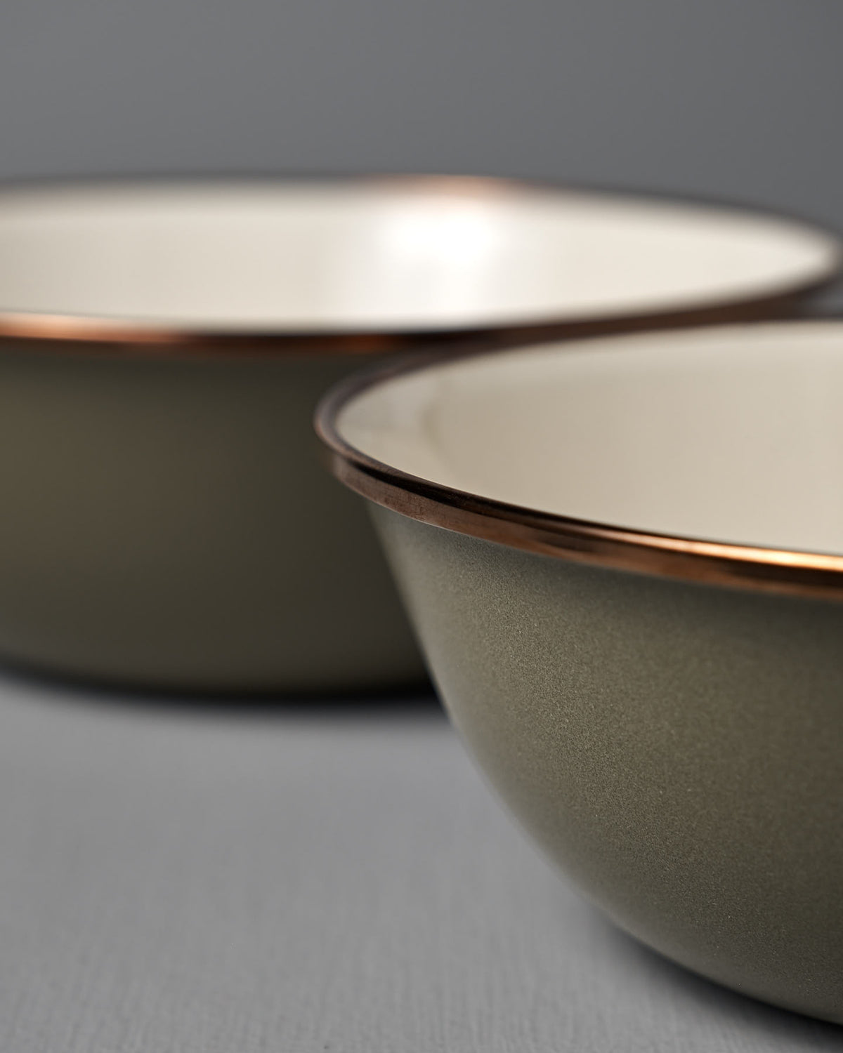 Close-up of Barebones Enamel Two-Tone Bowls in Olive Drab (Set of 2) with matte olive exteriors, glossy white interiors, metallic rims, and a vintage-inspired design, displayed against a neutral background.