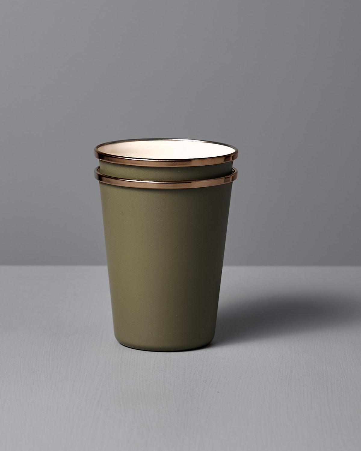 A set of two Barebones Enamel Two-Tone Tall Cups in Olive Drab, with a vintage look and rust-resistant finish, are stacked on a light gray surface against a plain gray background.