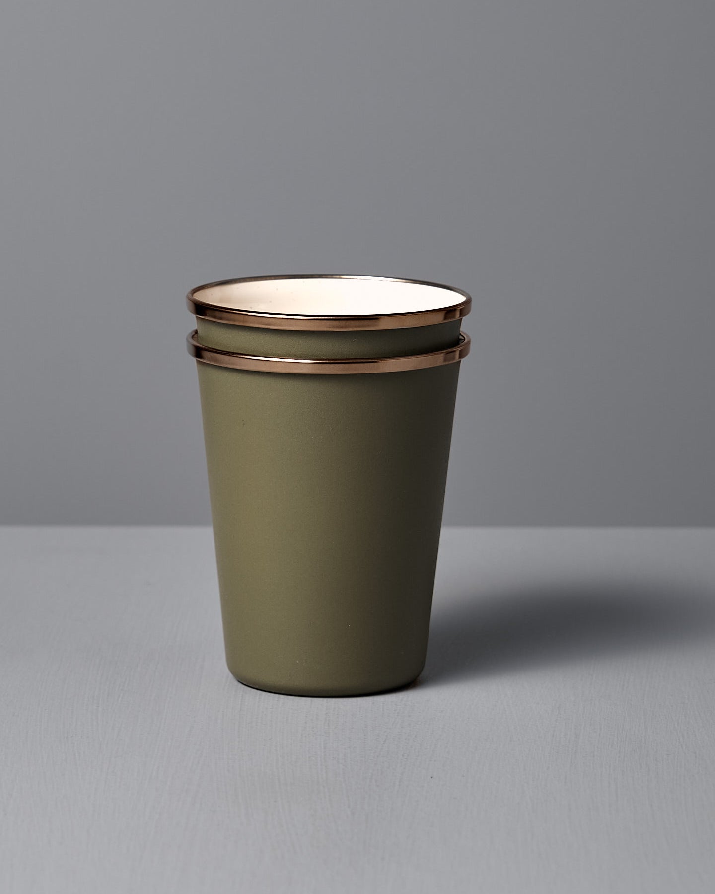 A set of two Barebones Enamel Two-Tone Tall Cups in Olive Drab, each with a stainless steel rim—one upright and one on its side—sit on a light gray surface against a gray background.