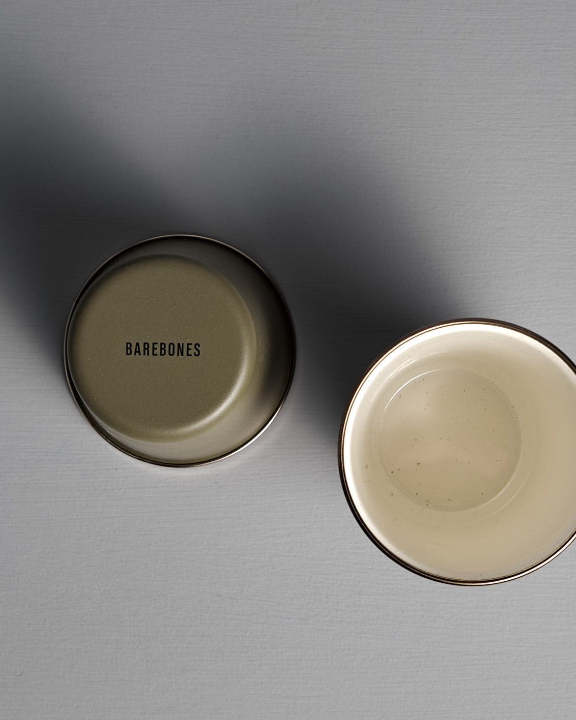 Two Barebones Enamel Two-Tone Tall Cups in Olive Drab are displayed—one upright and one inverted showing the brand logo—on a plain gray surface.