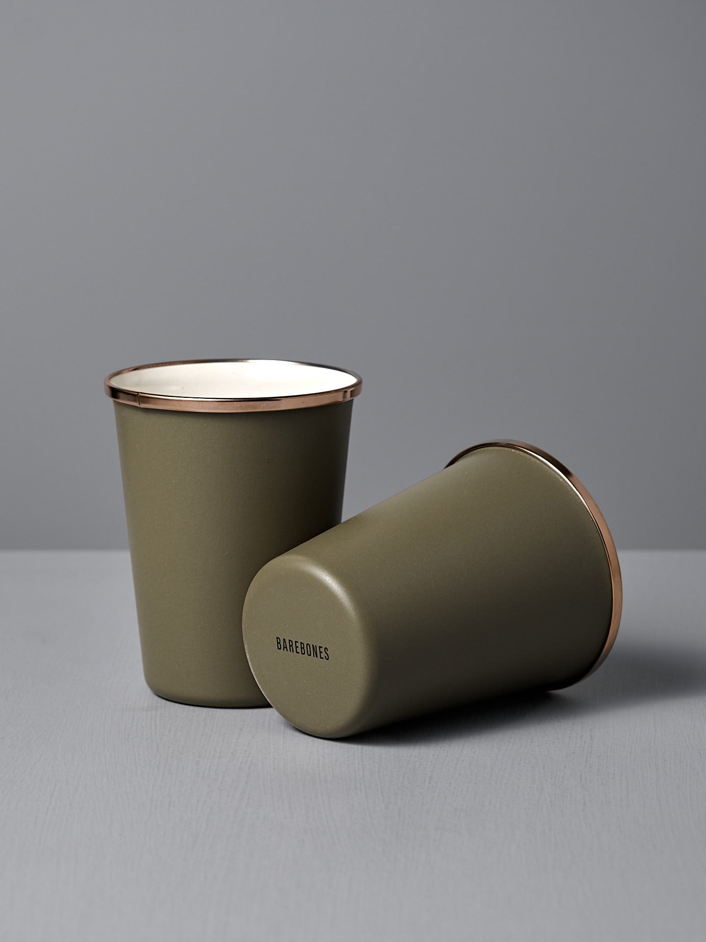 A set of two Barebones Enamel Two-Tone Tall Cups in Olive Drab, each with a stainless steel rim—one upright and one on its side—sit on a light gray surface against a gray background.