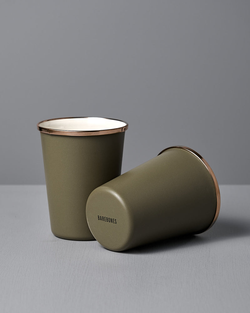 A set of two Barebones Enamel Two-Tone Tall Cups in Olive Drab, each with a stainless steel rim—one upright and one on its side—sit on a light gray surface against a gray background.