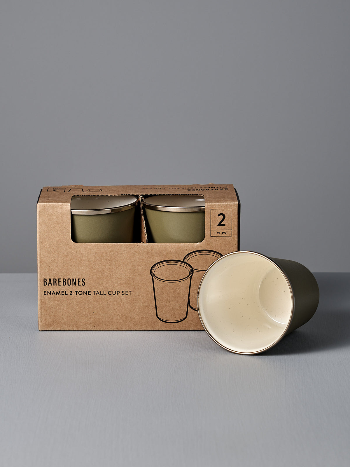 A boxed set of two Barebones Enamel Two-Tone Tall Cups in Olive Drab, with one cup outside the box displaying its cream interior, is shown against a plain grey background.