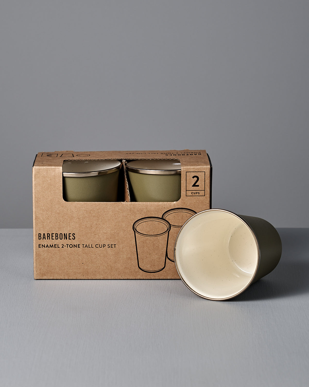 A boxed set of two Barebones Enamel Two-Tone Tall Cups in Olive Drab, with one cup outside the box displaying its cream interior, is shown against a plain grey background.