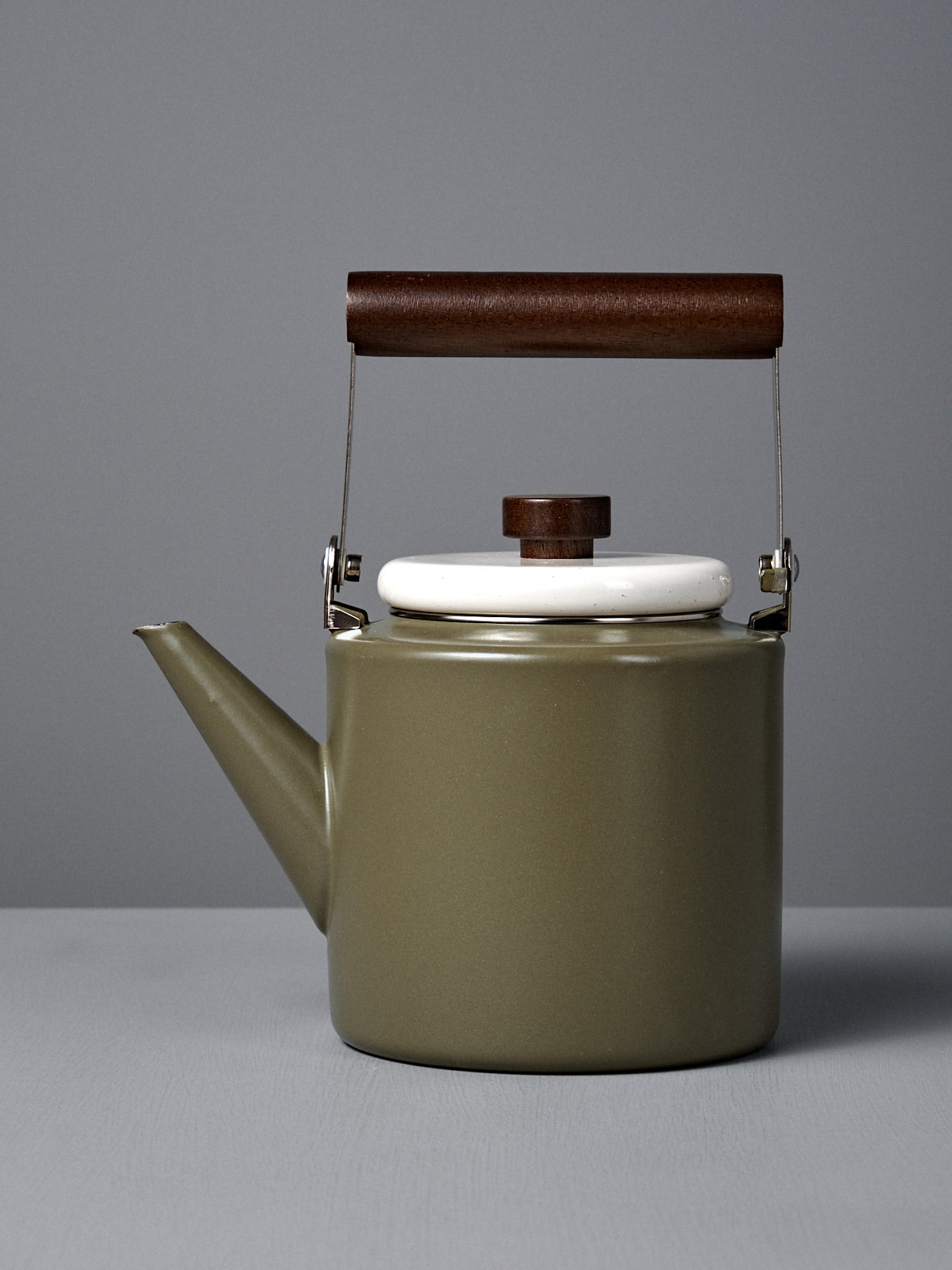 The Enamel Two-Tone Kettle in Olive Drab, featuring a white lid and dark walnut handle, is displayed on a plain gray surface with a gray background.