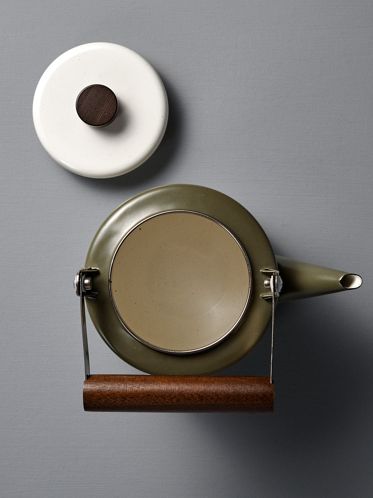 The Enamel Two-Tone Kettle - Olive Drab, featuring a walnut handle, is pictured from above beside its white lid with a dark wooden knob on a gray surface.