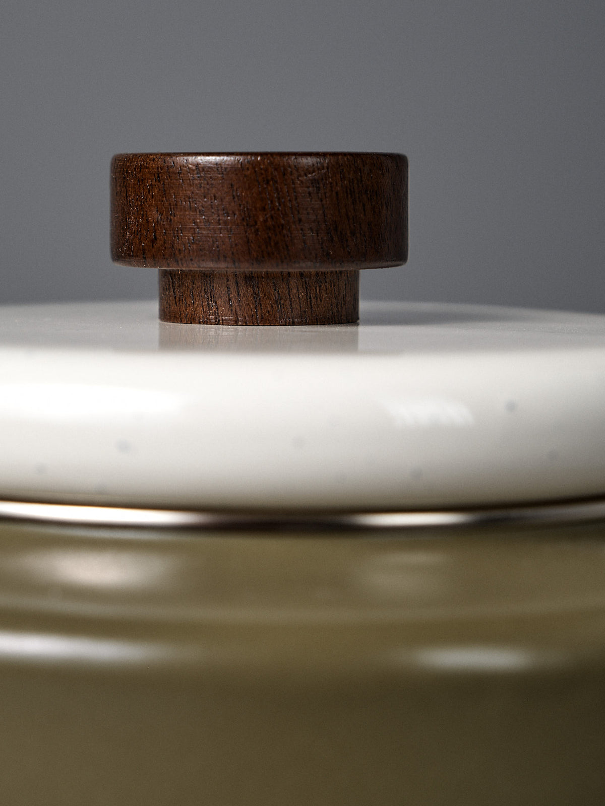 Close-up of the Enamel Two-Tone Kettle in Olive Drab, featuring a ceramic container with a white lid and a round dark wooden knob, set against a plain gray background.