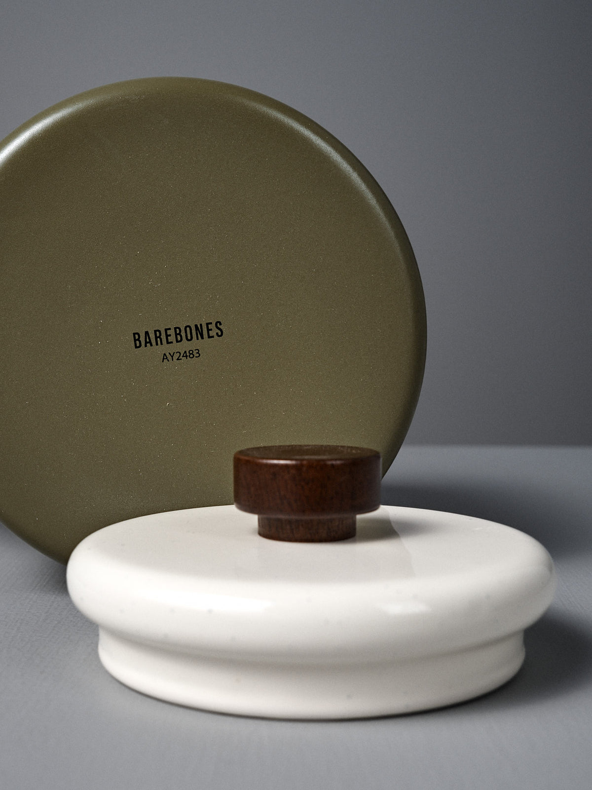 A round olive-green tin labeled &quot;Barebones&quot; stands upright behind the Enamel Two-Tone Kettle - Olive Drab, a flat, white ceramic container with a small wooden knob lid, evoking vintage-inspired charm.