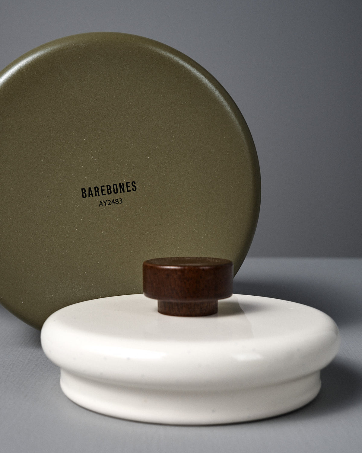 A round olive-green lid labeled "Barebones AY2483" stands upright behind the Enamel Two-Tone Kettle – Olive Drab's white enamelware lid with a dark walnut handle, both set on a neutral surface.