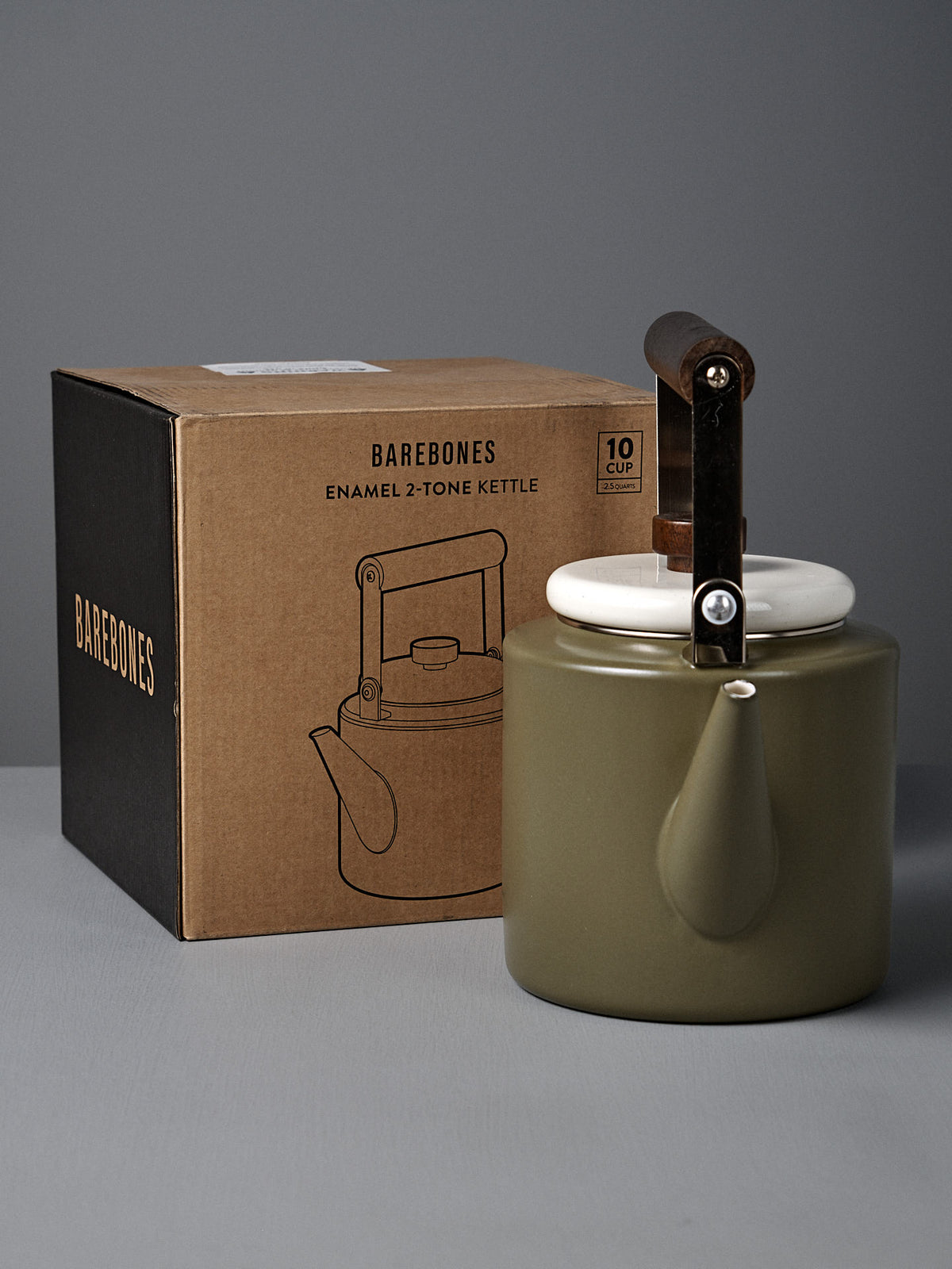 The Enamel Two-Tone Kettle - Olive Drab, featuring a vintage design with a walnut handle in green and white, sits next to its brown Barebones box on a gray surface.