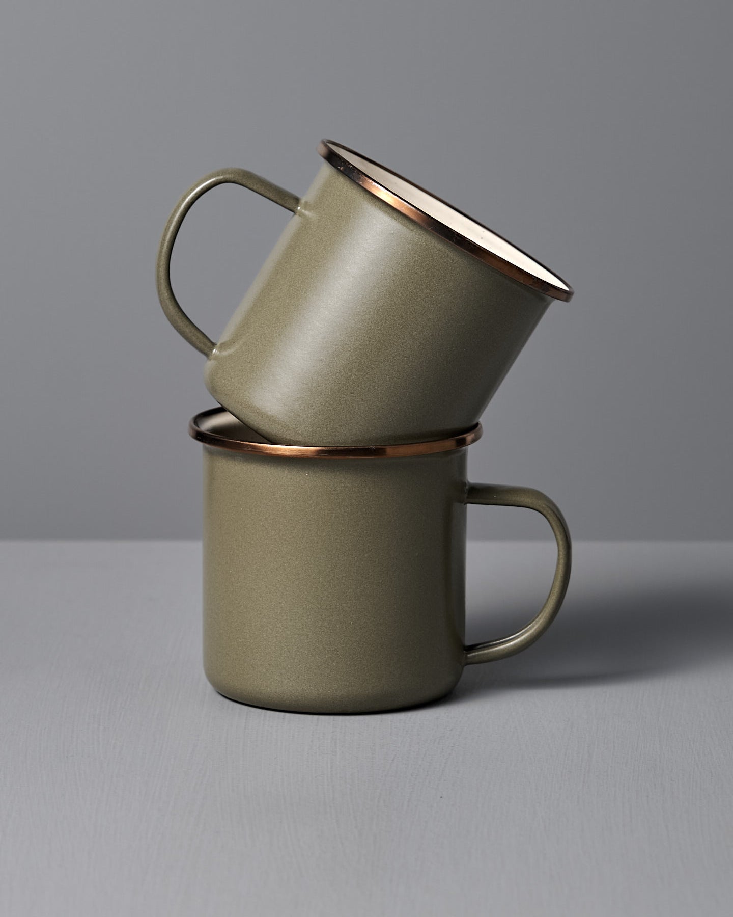 Two Barebones Enamel Two-Tone Mugs in Olive Drab (Set of 2) are stacked together against a neutral gray backdrop, highlighting their vintage-inspired design and sturdy handles.