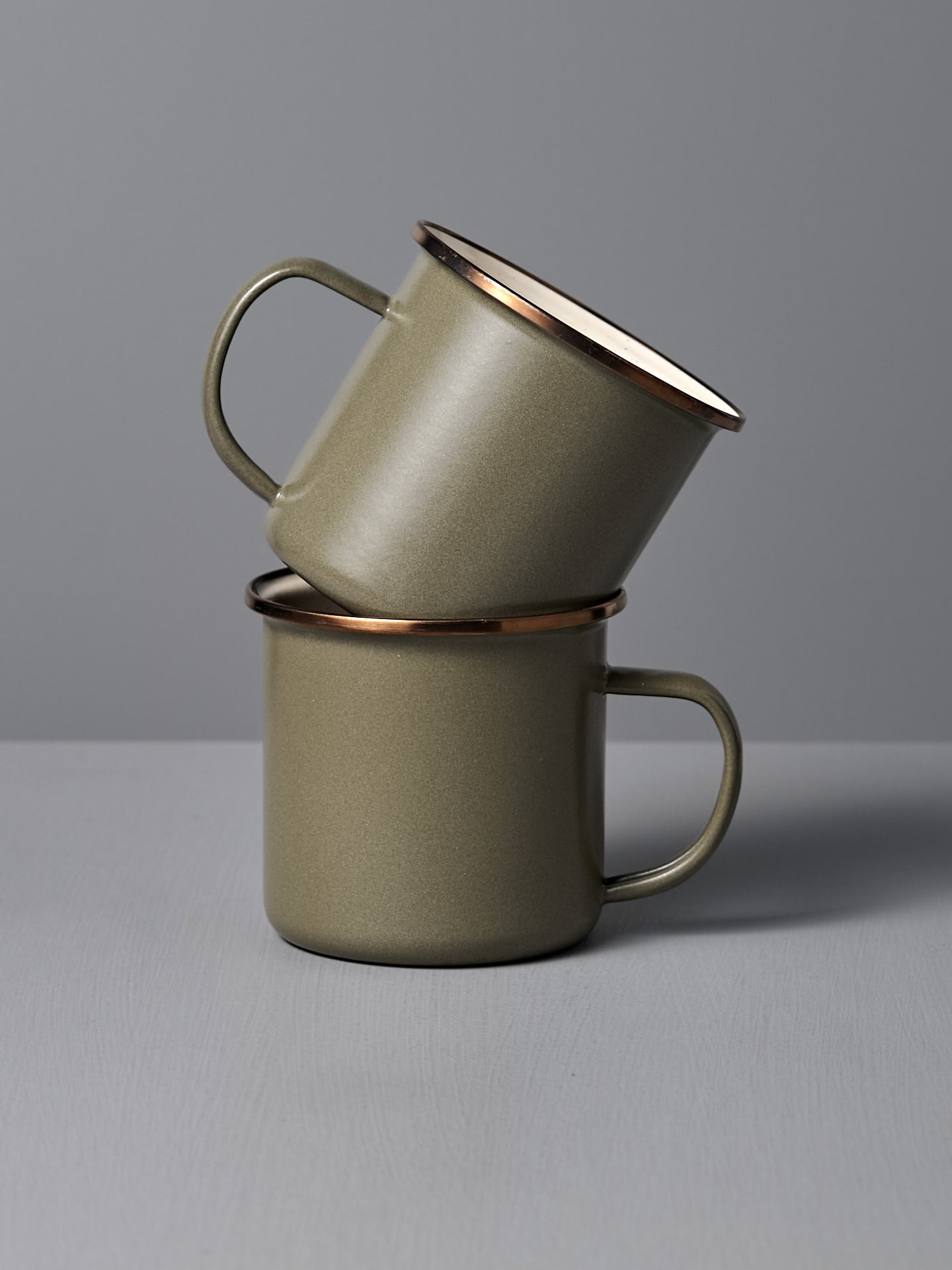 Two Barebones Enamel Two-Tone Mugs in Olive Drab (Set of 2) are stacked together against a neutral gray backdrop, highlighting their vintage-inspired design and sturdy handles.