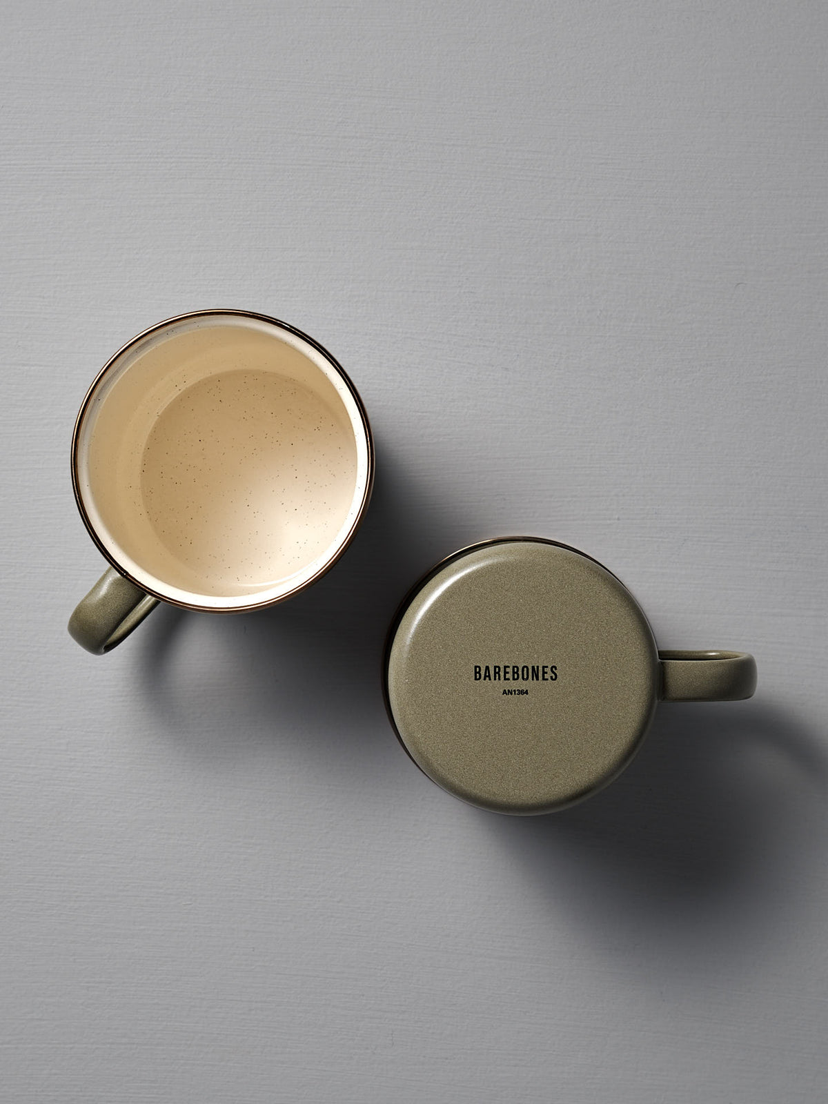 Two Barebones Enamel Two-Tone Mugs in Olive Drab rest on a gray surface—one upright with its interior visible, the other upside down showing the &quot;Barebones&quot; logo on its base.