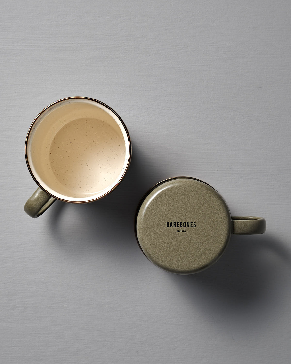 Two Barebones Enamel Two-Tone Mugs in Olive Drab rest on a gray surface—one upright with its interior visible, the other upside down showing the "Barebones" logo on its base.