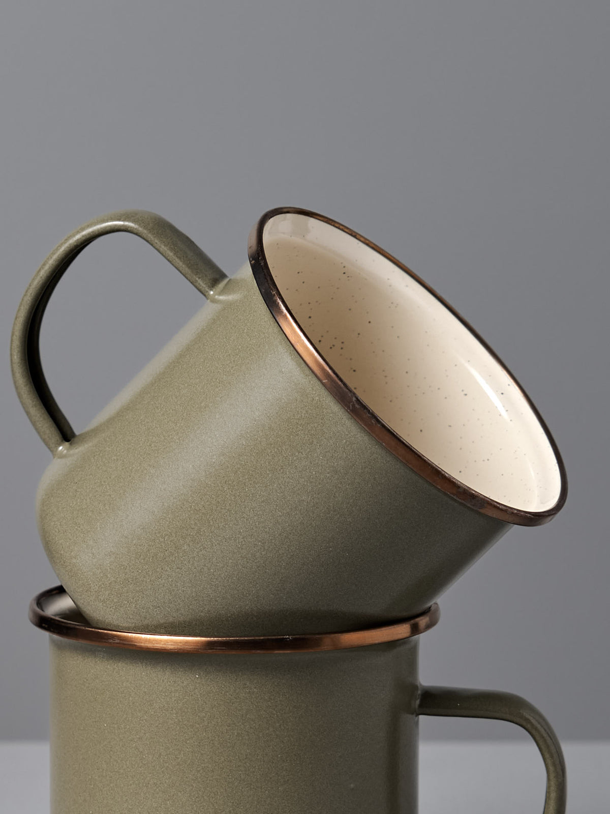 The Barebones Enamel Two-Tone Mug – Olive Drab (Set of 2) features vintage-inspired olive green mugs with brown rims, shown stacked against a plain gray background.