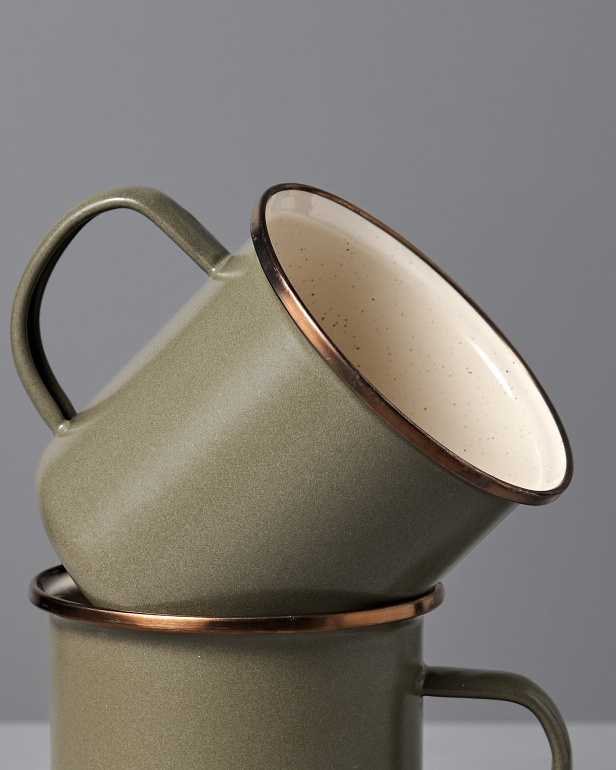 The Barebones Enamel Two-Tone Mug – Olive Drab (Set of 2) features vintage-inspired olive green mugs with brown rims, shown stacked against a plain gray background.