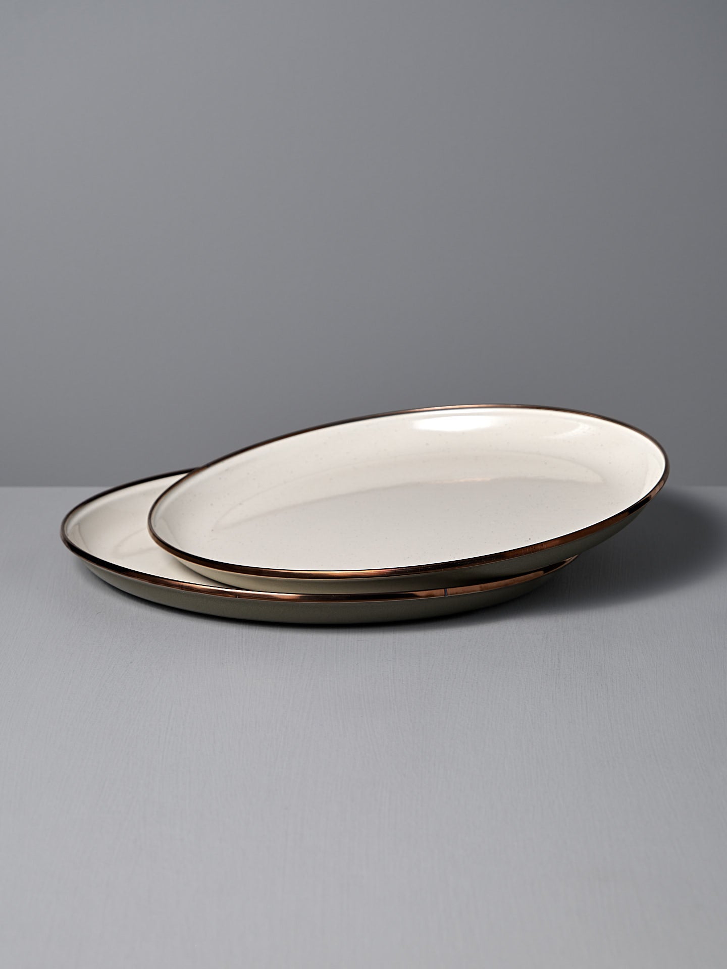 The Barebones Enamel Two-Tone Deep Plates in Olive Drab (Set of 2) are stacked on a light grey surface, showcasing their vintage-inspired design and brown rims against a plain grey background.