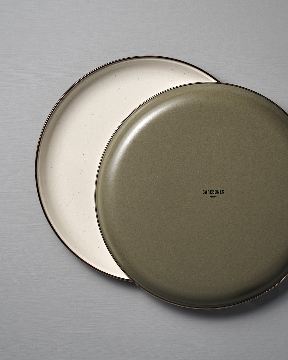 Two Barebones Enamel Two-Tone Deep Plates in Olive Drab overlap on a light gray surface, highlighting their vintage-inspired look that complements enamelware cups. This set includes two plates featuring the Barebones brand.