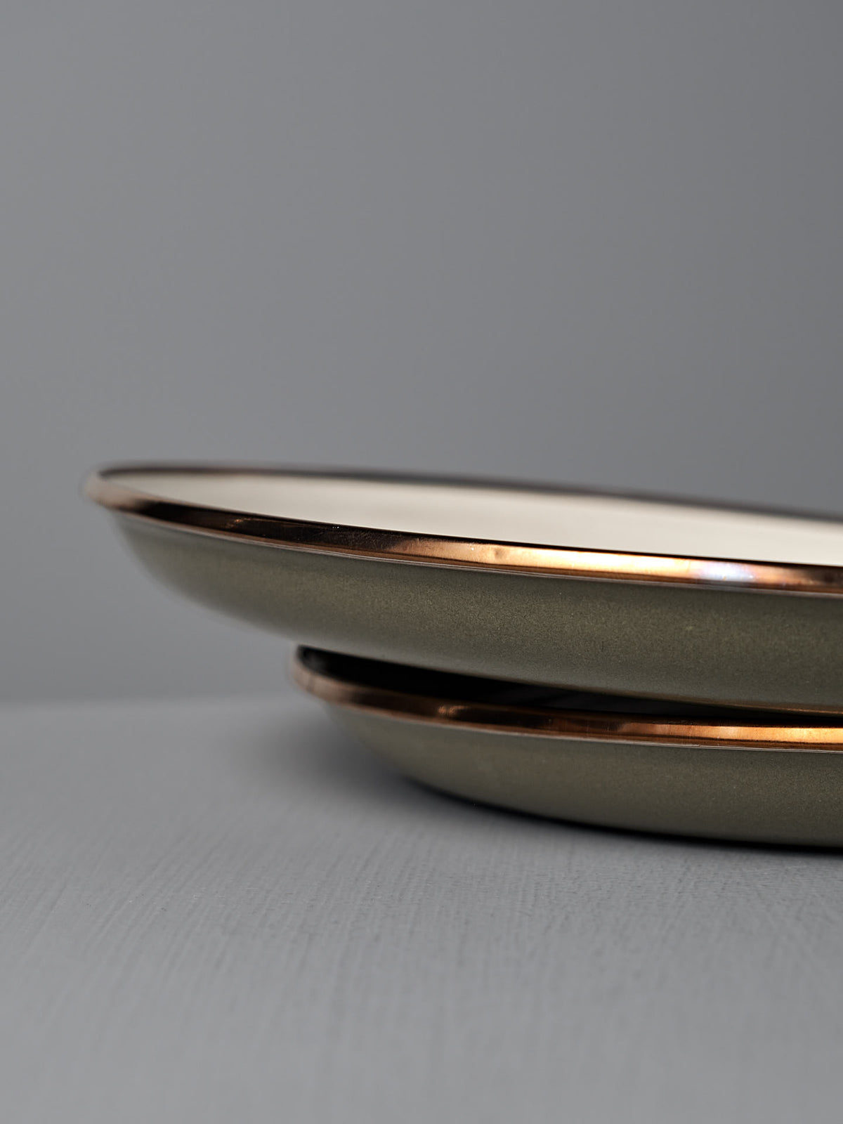 A set of two Barebones Enamel Two-Tone Deep Plates in Olive Drab are stacked on a gray surface, their vintage-inspired metallic rims highlighted against a plain gray background.