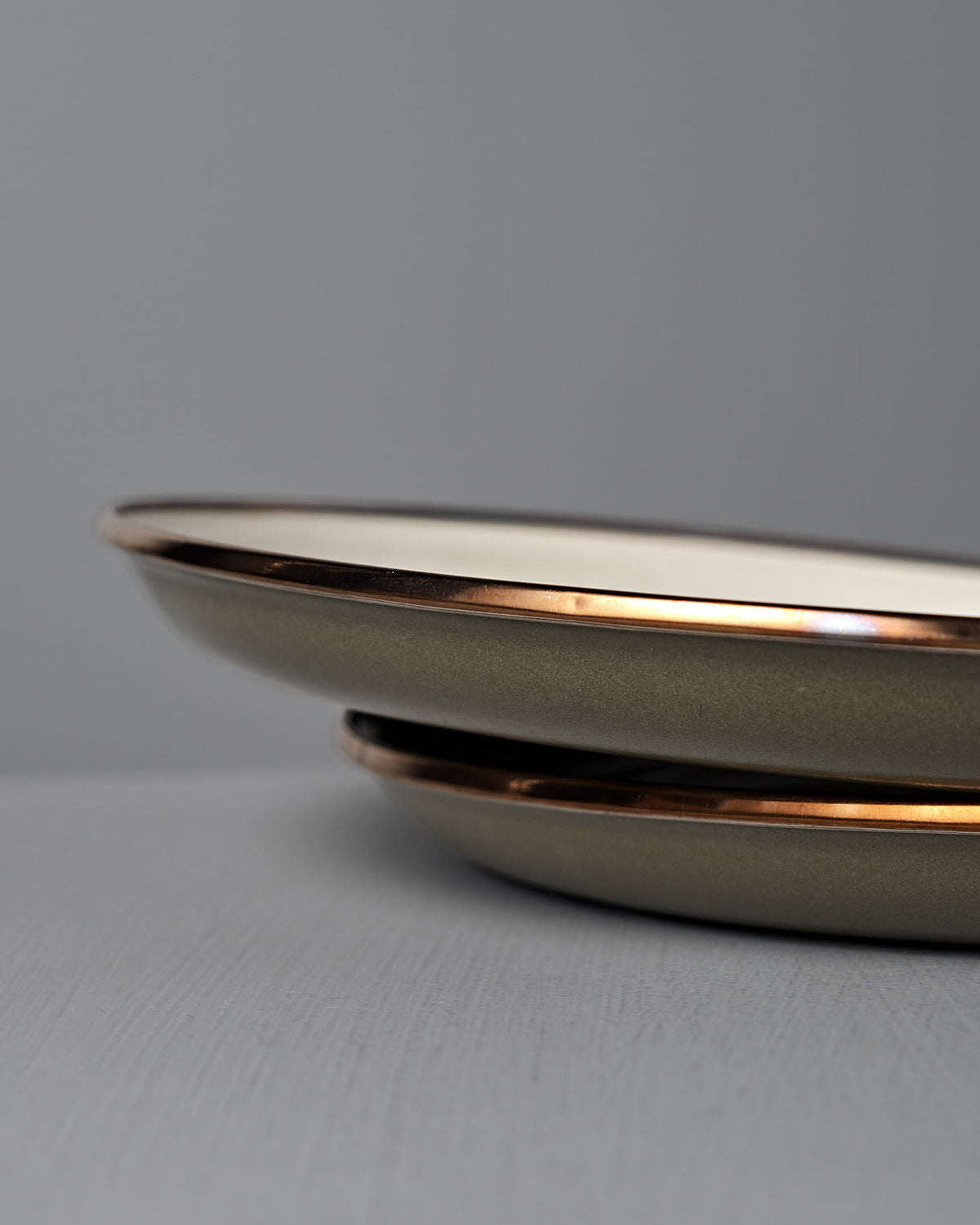 A set of two Barebones Enamel Two-Tone Deep Plates in Olive Drab are stacked on a gray surface, their vintage-inspired metallic rims highlighted against a plain gray background.