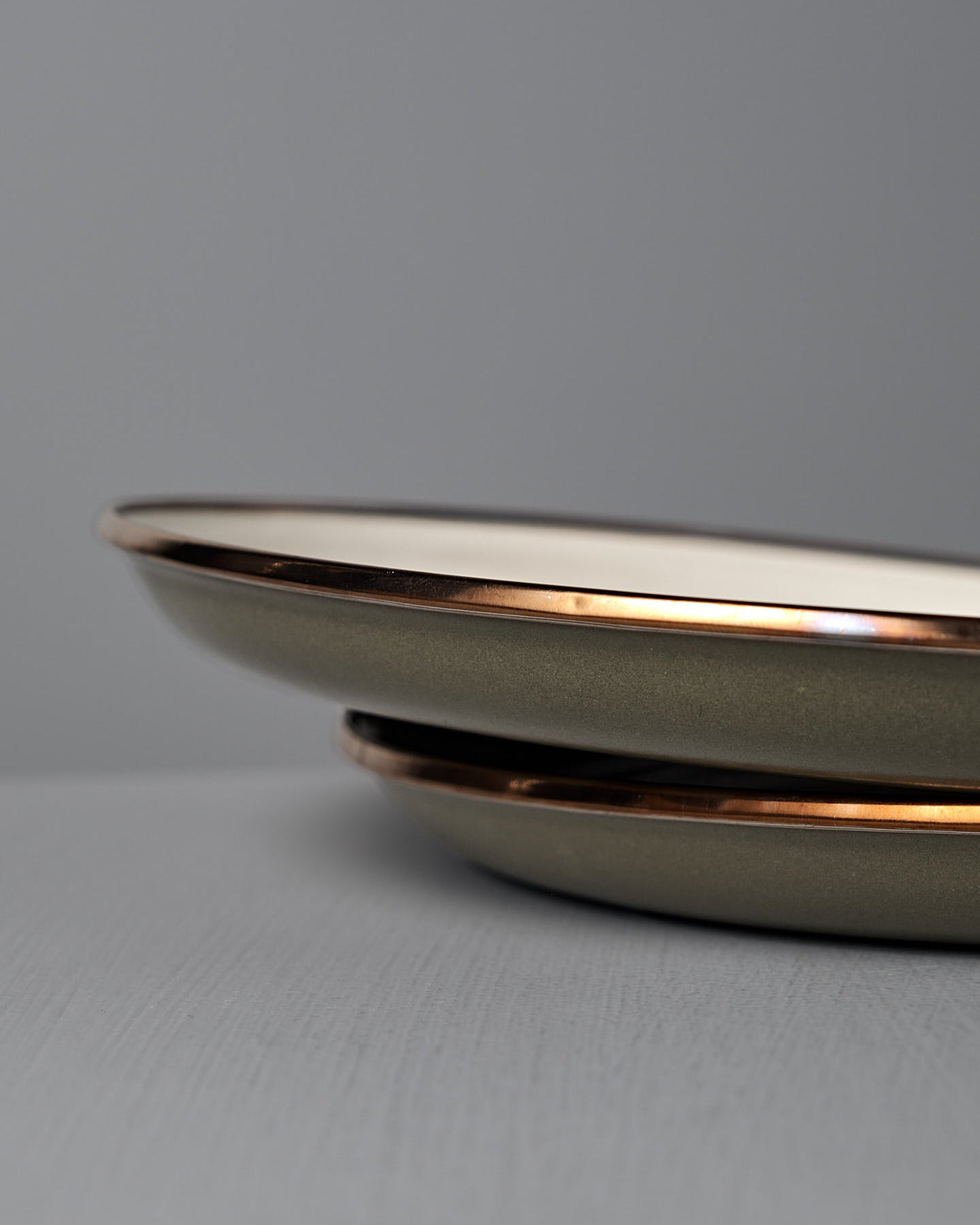 The Barebones Enamel Two-Tone Deep Plates in Olive Drab (Set of 2) are stacked on a light grey surface, showcasing their vintage-inspired design and brown rims against a plain grey background.