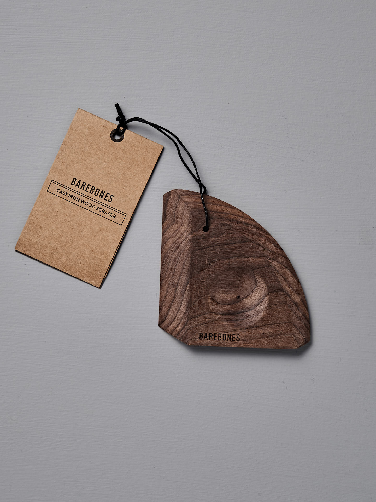 The Barebones Cast Iron Wood Scraper, an ergonomic tool for cleaning cookware, features a branded tag attached by a black string and is shown on a gray surface.