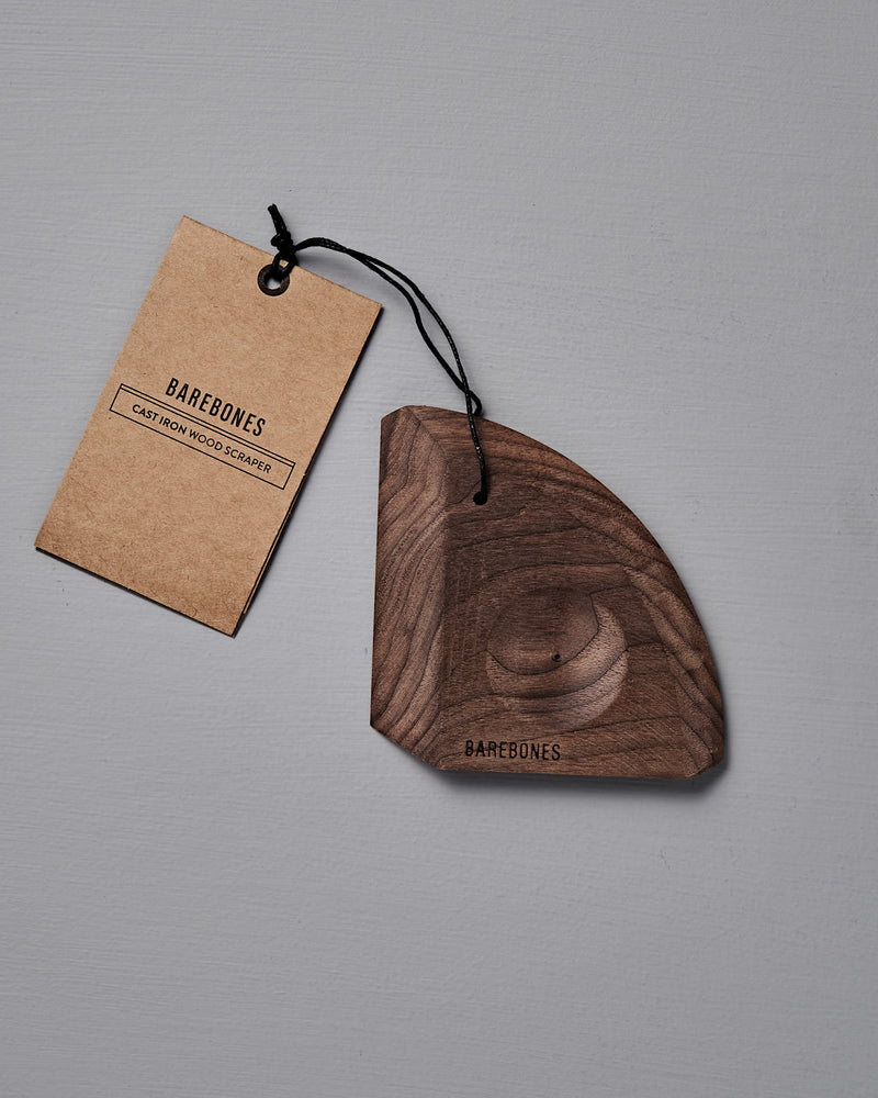 The Barebones Cast Iron Wood Scraper, an ergonomic tool for cleaning cookware, features a branded tag attached by a black string and is shown on a gray surface.