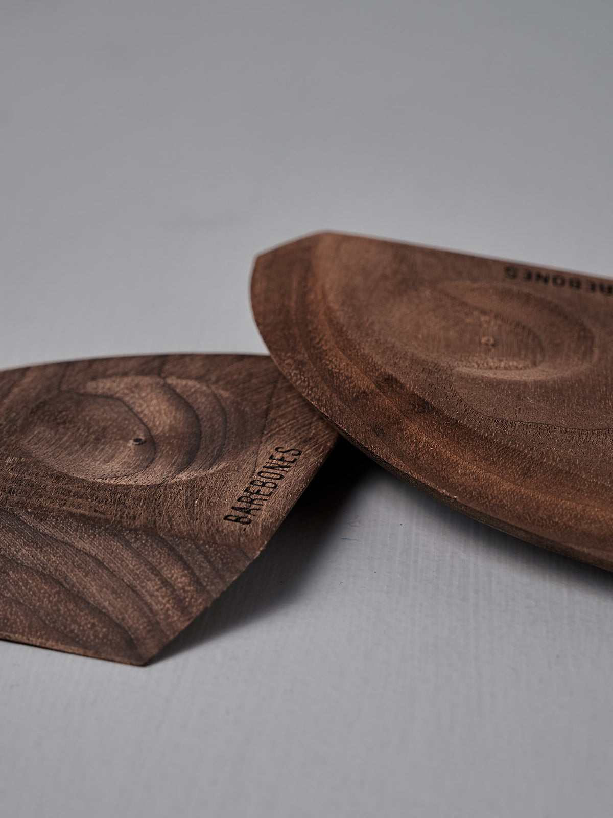 Two Barebones Cast Iron Wood Scrapers, featuring carved walnut with visible grain and embossed branding, rest on a smooth light gray surface—an ergonomic kitchen tool perfect for cleaning cookware.