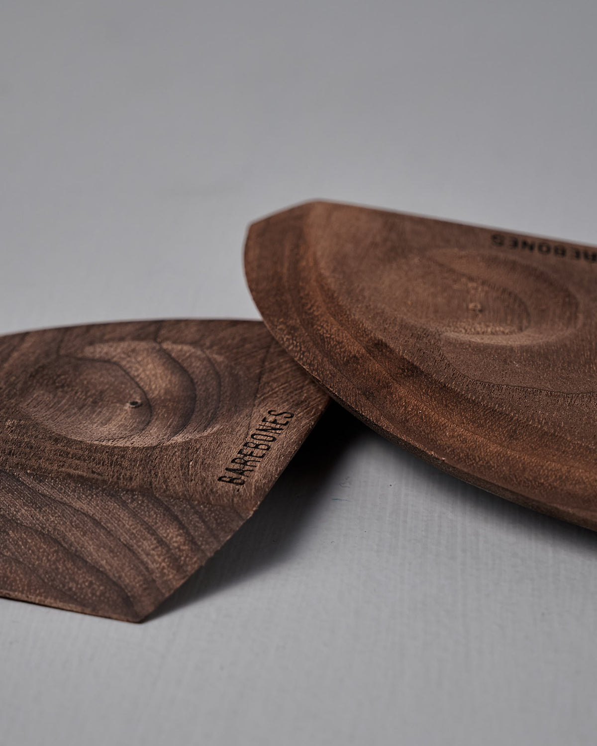 Two Barebones Cast Iron Wood Scrapers, featuring carved walnut with visible grain and embossed branding, rest on a smooth light gray surface—an ergonomic kitchen tool perfect for cleaning cookware.