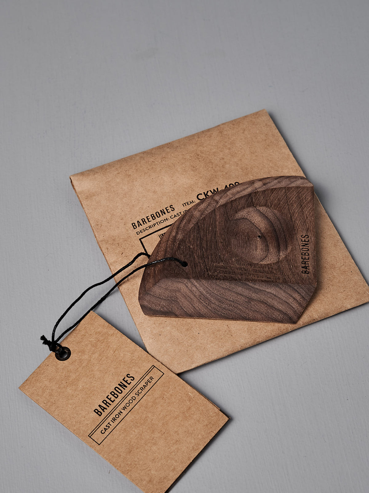 The Barebones Cast Iron Wood Scraper, an ergonomic kitchen tool for cleaning cookware, sits on a brown paper envelope next to a branded tag with black string.