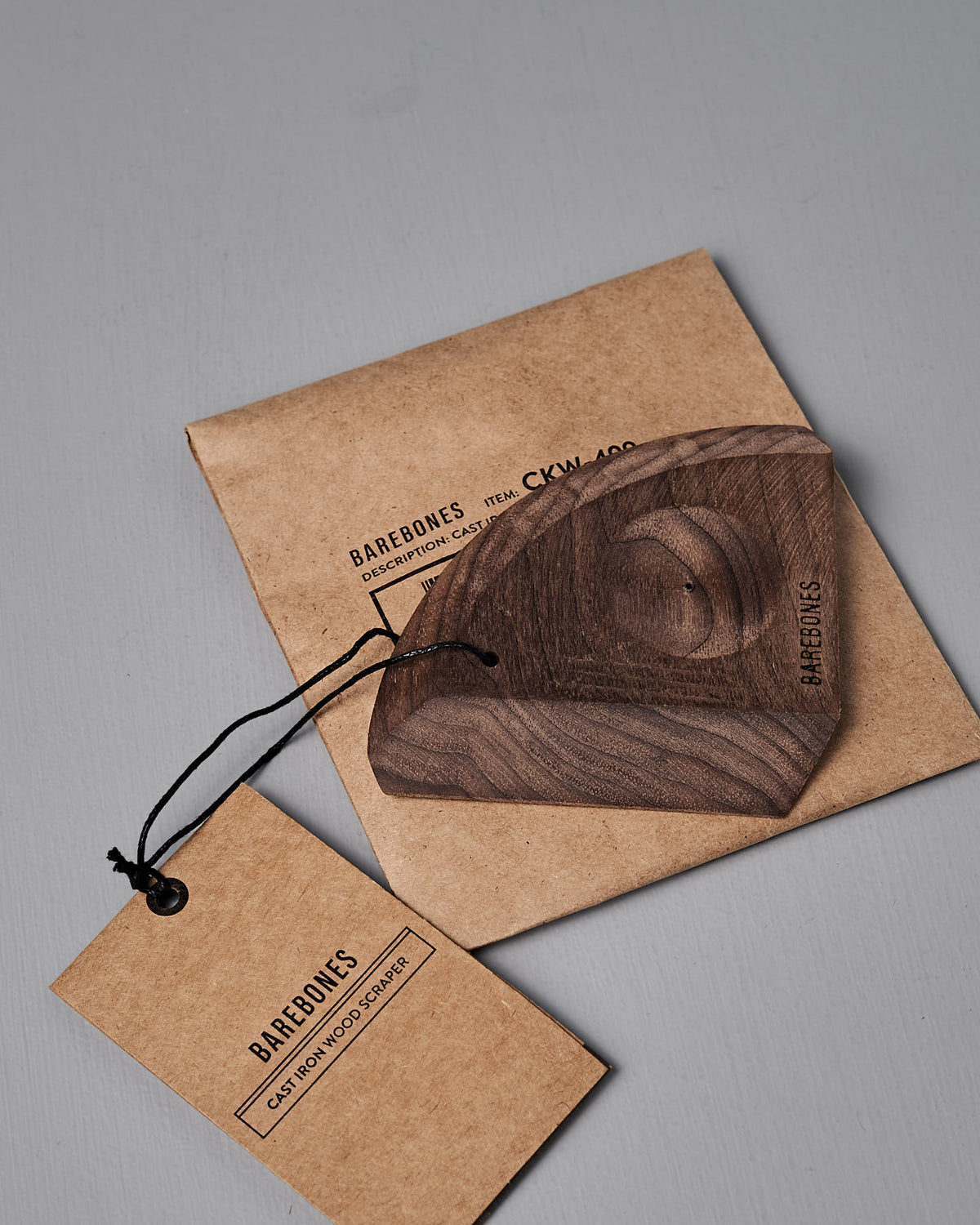The Barebones Cast Iron Wood Scraper, an ergonomic kitchen tool for cleaning cookware, sits on a brown paper envelope next to a branded tag with black string.