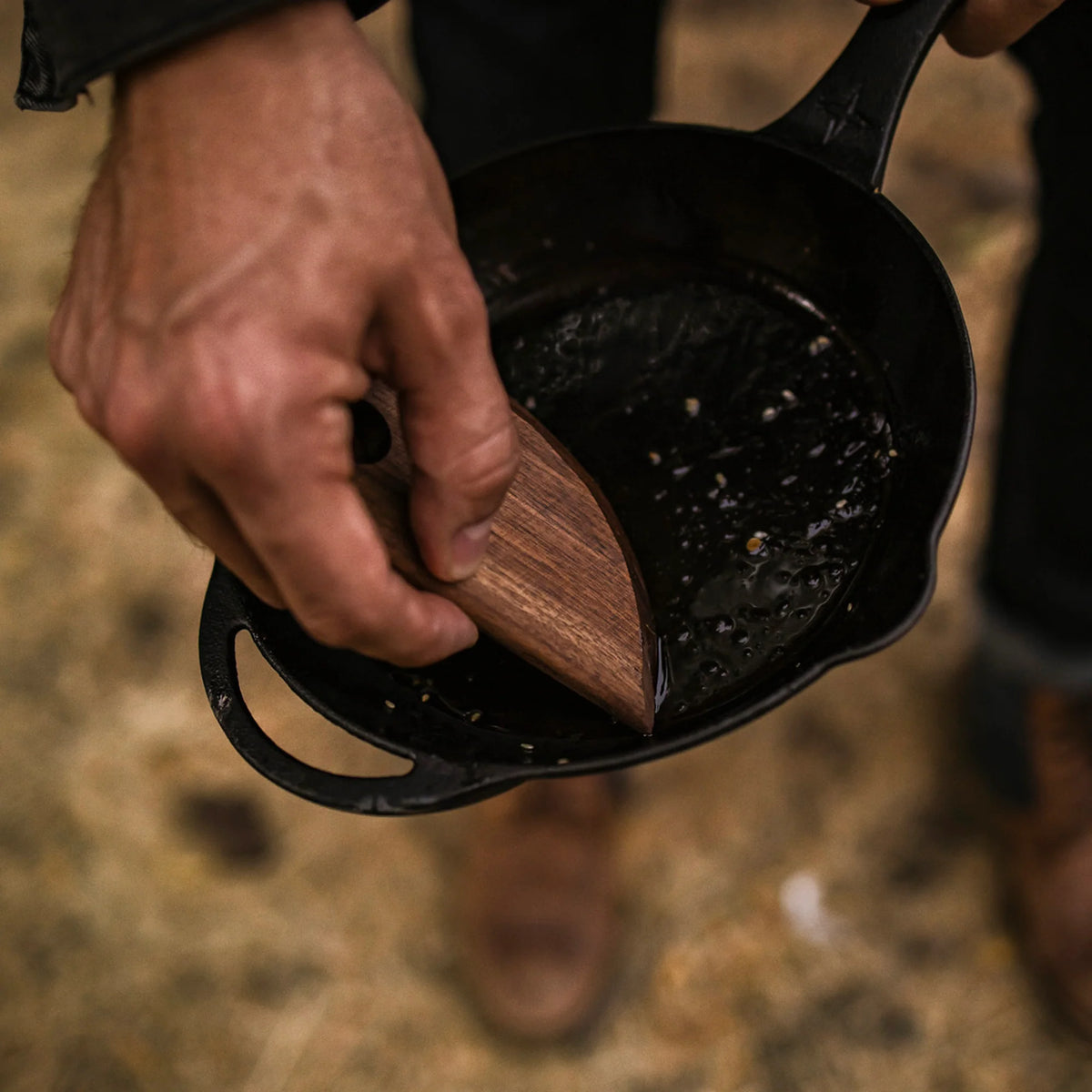 A person uses the Barebones Cast Iron Wood Scraper to remove food residue from a black cast iron skillet outdoors, showcasing effective cookware cleaning.